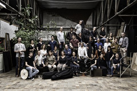 Eine große Gruppe von Menschen posiert gemeinsam in einem industriellen Raum. Einige halten Musikinstrumente, andere stehen lässig.  © Mirjam Devriendt