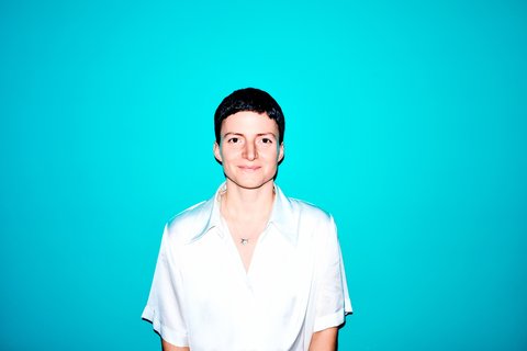 Person mit kurzer Frisur und weißem Hemd vor einem leuchtend blauen Hintergrund. Sie lächelt sanft in die Kamera.