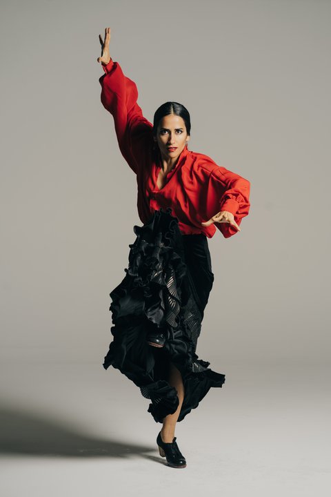 Frau in roter Bluse und schwarzem Rock tanzt leidenschaftlich Flamenco, eine Hand erhoben, in einem Studio vor neutralem Hintergrund.  © Tarantos Flamenco