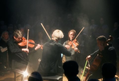 Ein Streichquartett spielt in einem abgedunkelten Raum. Das Publikum sitzt im Halbrund und lauscht aufmerksam dem Konzert.