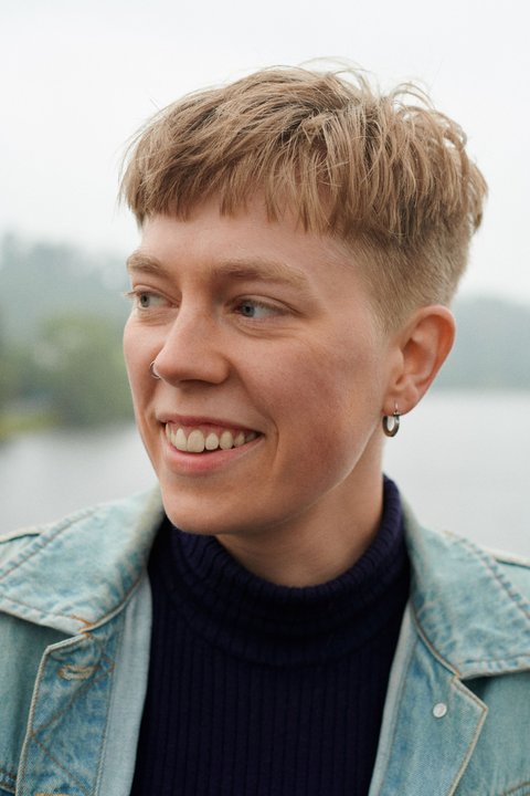 Eine Person posiert vor einem Fluss. Deren Haare sind kurz und dey lächelt. Deren Kopf ist von der Kamera weggedreht.